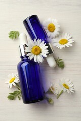 Bottles of essential oil and chamomile flowers on light wooden table, flat lay