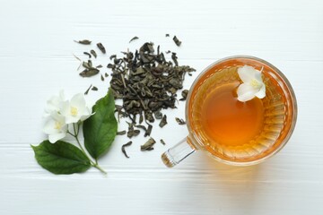 Aromatic jasmine tea in glass cup, flowers and leaves on white wooden table, flat lay