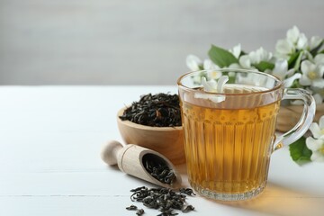 Aromatic jasmine tea in glass cup, flowers and dried leaves on white wooden table, closeup. Space...