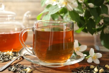 Aromatic jasmine tea in glass cup, teapot, brew and flowers on wooden table, closeup