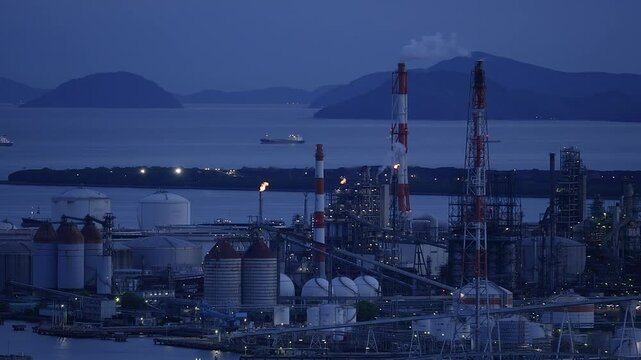 A sprawling industrial plant with smoking chimneys and burning flare stacks at dusk.