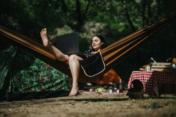 A young woman enjoys a moment of leisure in a forest campsite while working remotely. She sits comfortably in a hammock using her laptop, merging the outdoors with modern lifestyle and productivity.