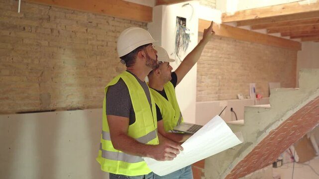 Video of Two male construction workers in hard hats and reflective vests examine blueprints and use a laptop at a building site