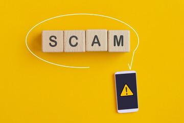 For investment scam, fraud and Ponzi scheme concept.  SCAM text on wooden cube blocks  and exclamation mark on mobile phone with yellow background