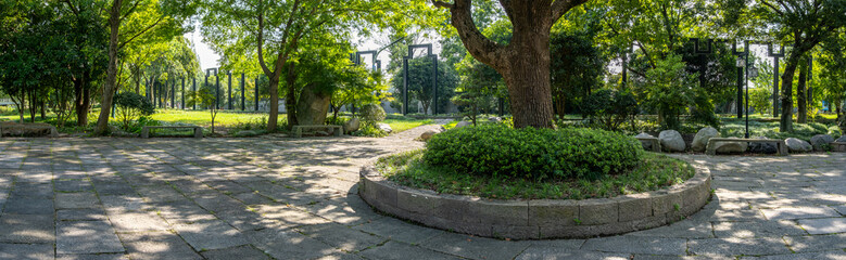 Panoramic view of an outdoor public plaza shaded by mature trees, with circular planters, stone...