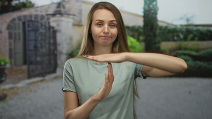 Woman making timeout gesture outdoors in garden setting with relaxed expression and light clothing, surrounded by lush greenery and classic architecture, serene afternoon ambiance.
