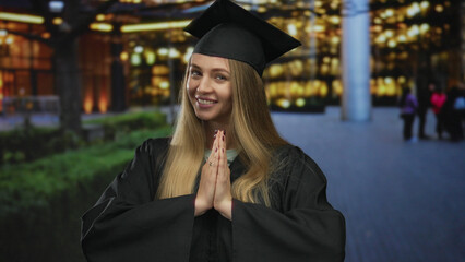 Young woman in graduation attire standing on a city street at night, with hands in a gratitude...