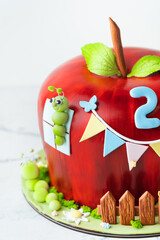 Red apple shaped fondant cake decorated with mastic edible caterpillar, green leaves and moss. Orchard fruits themed birthday cake isolated on white background