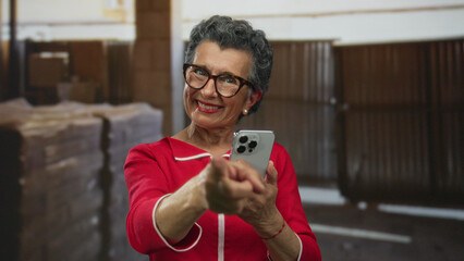 Senior woman with grey hair and glasses, smiling and pointing smartphone in construction site...