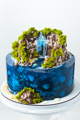 Beautiful birthday cake in the shape of an island decorated with chocolate edible mountains, greenery with waterfall. Tropical island cake surrounded with blue jelly ocean water