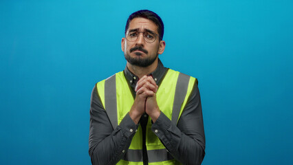 Man wearing a reflective vest and glasses stands against a blue background appearing thoughtful and...