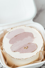 Small bento cake with white cream cheese frosting decorated with gold painting and pastel cream smears. Beautiful anniversary cake on white marble background