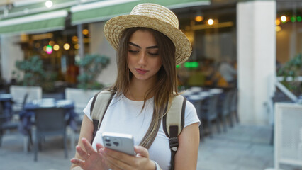 Woman standing on busy city street using smartphone with straw hat and backpack, blending urban exploration with digital connectivity amidst exterior daytime vibrancy.