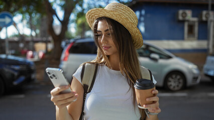 Woman in straw hat holding coffee cup and smartphone on urban street with cars and trees.