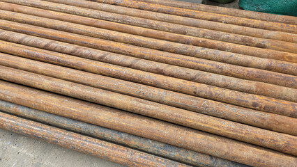Horizontal photo of multiple old, corroded steel pipes with surface rust, stored outdoors on a concrete surface for recycling or reuse
