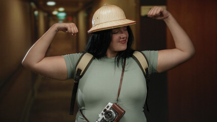 Woman tourist flexing in a hotel hallway with a camera around her neck, wearing a safari hat,...
