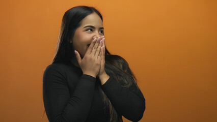 Woman laughing with hands over mouth standing against a vibrant orange wall, expressing joy and...