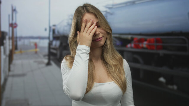 Woman with blonde hair hand covering forehead on urban sidewalk next to parked truck ; exasperation frustration.