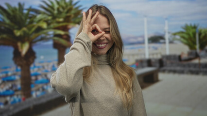 Blonde woman makes ok sign with hand around eye at beach with palm trees and umbrellas behind;...