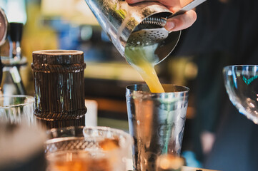 Bartender pouring yellow cocktail from shaker into metal cup on bar counter with tools and...
