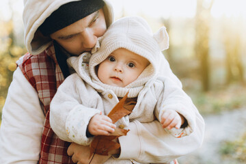 Happy father hugging and kissing cute baby son in soft evening light in autumn woods. Fatherhood. Stylish man with adorable child in cozy warm sweaters embracing in fall park