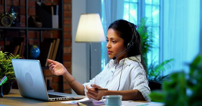 Businesswoman using headset for call at computer desk in modern cozy office, Indian Asian female attending teleconference with microphone headphones working on laptop communicating professionally