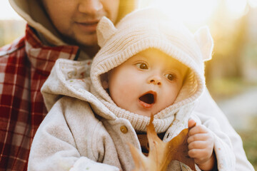 Happy cute baby son holding autumn leaf in fathers hands in soft evening light in autumn woods. Fatherhood. Stylish man with adorable child in cozy warm sweaters hugging in fall park