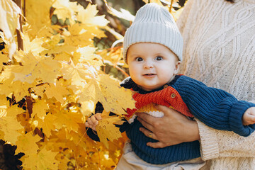 Cute baby boy with autumn yellow leaves close up. Childhood. Stylish mother holding adorable son in cozy warm sweaters in fall park