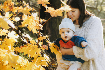 Happy mother hugging cute baby son in autumn woods with yellow leaves. Motherhood. Stylish woman with adorable child in cozy warm sweaters relaxing in fall park