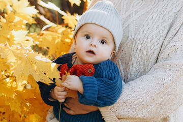 Cute baby boy in mothers hands close up in autumn yellow leaves. Childhood. Stylish woman with adorable son in cozy warm sweaters relaxing in fall park