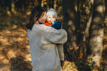 Happy mother hugging cute baby son in autumn woods. Motherhood. Stylish woman with adorable child in cozy warm sweaters embracing and relaxing in fall park