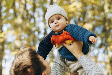 Cute baby son with funny emotions in father hands close up in autumn woods. Fatherhood. Stylish man with adorable child in cozy warm sweaters having fun and relaxing in fall park