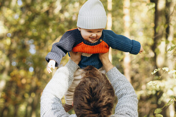 Cute baby son with funny emotions in father hands close up in autumn woods. Fatherhood. Stylish man with adorable child in cozy warm sweaters having fun and relaxing in fall park