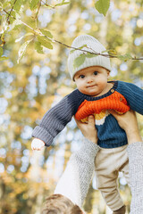 Cute baby son with funny emotions in father hands close up in autumn woods. Fatherhood. Stylish man with adorable child in cozy warm sweaters having fun and relaxing in fall park