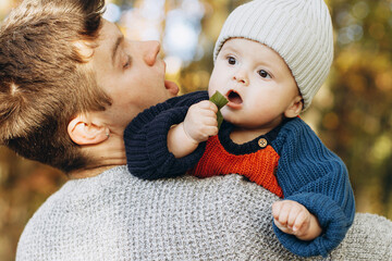 Cute baby son with funny emotions holding leaves close up in father hands  in autumn woods. Fatherhood. Stylish man with adorable child in cozy warm sweaters relaxing in fall park