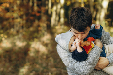 Happy father hugging and kissing cute baby son in autumn woods. Fatherhood. Stylish man with adorable child in cozy warm sweaters embracing and relaxing in fall park