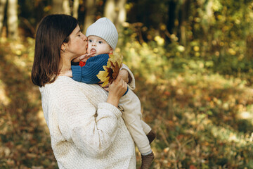 Happy mother hugging and kissing cute baby son in autumn woods. Motherhood. Stylish woman with adorable child in cozy warm sweaters relaxing in fall park