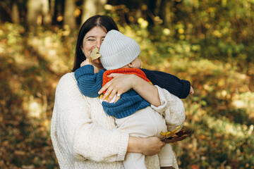 Happy mother hugging cute baby son in autumn woods. Motherhood. Stylish woman with adorable child in cozy warm sweaters embracing and relaxing in fall park