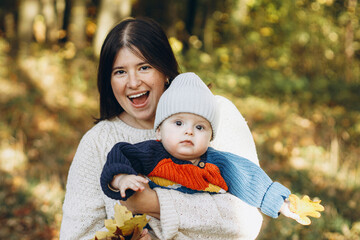 Happy mother hugging cute baby son in autumn woods. Motherhood. Stylish woman with adorable child in cozy warm sweaters embracing and relaxing in fall park
