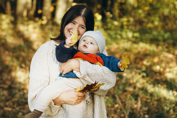 Happy mother hugging cute baby son in autumn woods. Motherhood. Stylish woman with adorable child in cozy warm sweaters embracing and relaxing in fall park