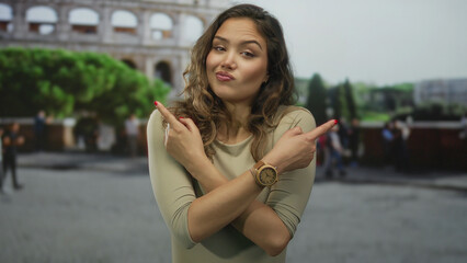 Fototapeta premium Hispanic woman crosses arms and points finger to both sides at coliseum building; confusion dilemma.