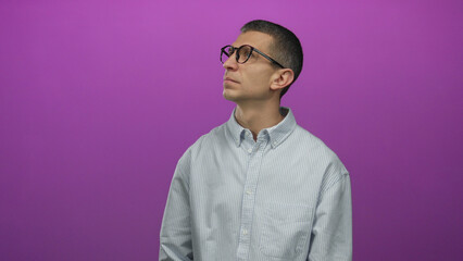 Man in glasses and striped shirt looks up against a purple wall, isolated from the background,...