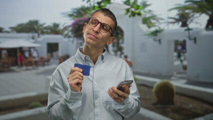 Caucasian man holding credit card and smartphone standing outdoors on street with relaxed...