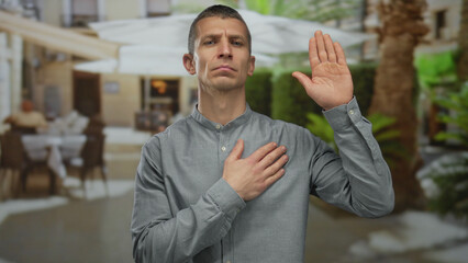 Man making a pledge gesture outdoors with a serious expression, in a street setting featuring...