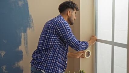 Young man with beard using masking tape on window frame in living room during home renovation...