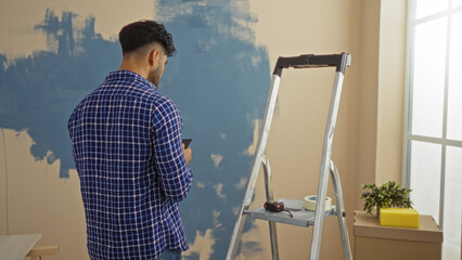 Young man with beard using mobile in living room near ladder and paint supplies, wearing plaid...