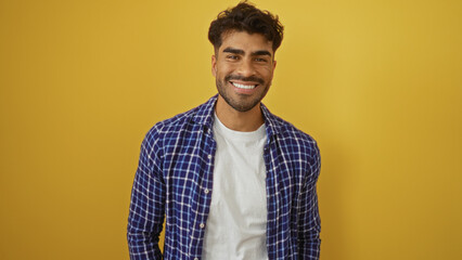 Young hispanic man with a beard smiling against an isolated yellow background, exuding attractiveness and confidence in casual attire.