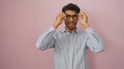 Young man with beard adjusts glasses while standing against a pink wall, exuding confidence and...