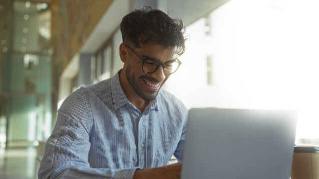 Young man with beard working on laptop in modern hospital office, wearing glasses, showing happiness and focus in bright indoor setting. - Powered by Adobe