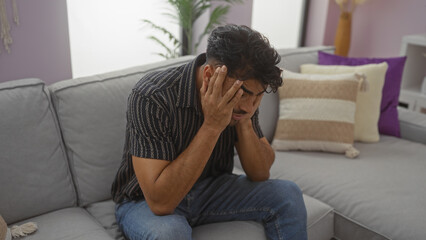 Young man with beard sitting on sofa in modern living room holding head looking stressed and thoughtful